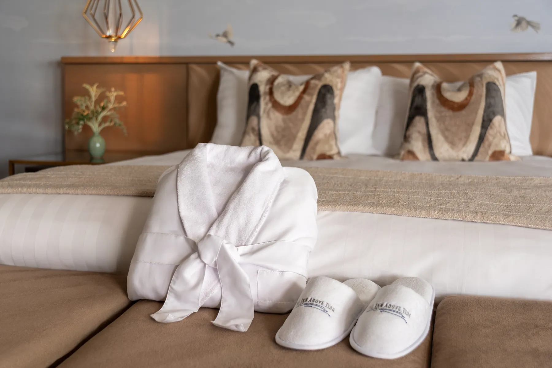 A luxury plush robe and white fluffy slippers sit at the foot of a neatly made bed.