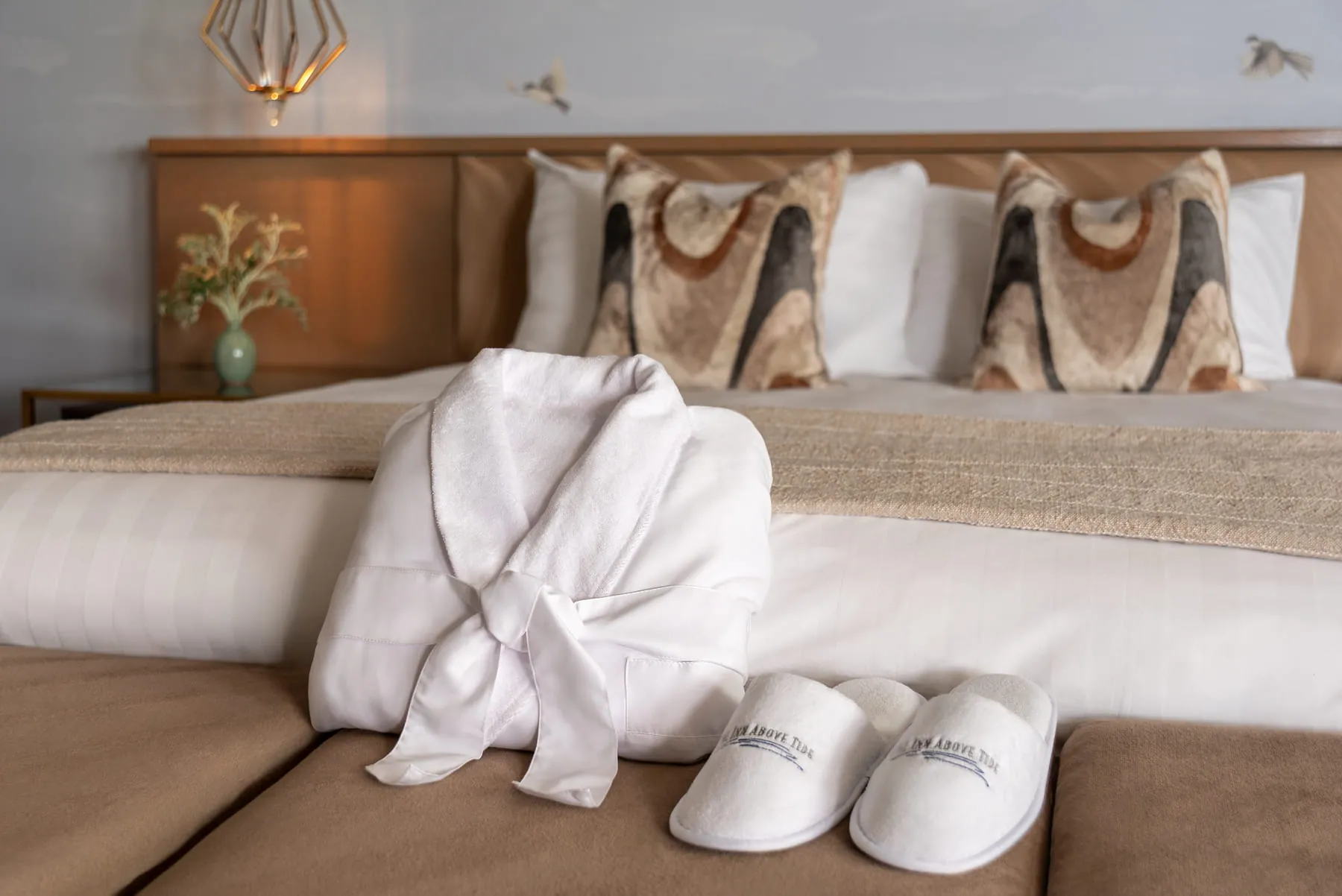 White robe and Inn Above Tide slippers on a bed with patterned pillows